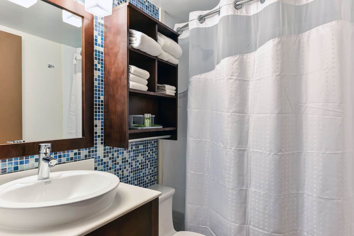A bathroom with a white sink, a mirror, a shower curtain, and a shelf with folded towels.