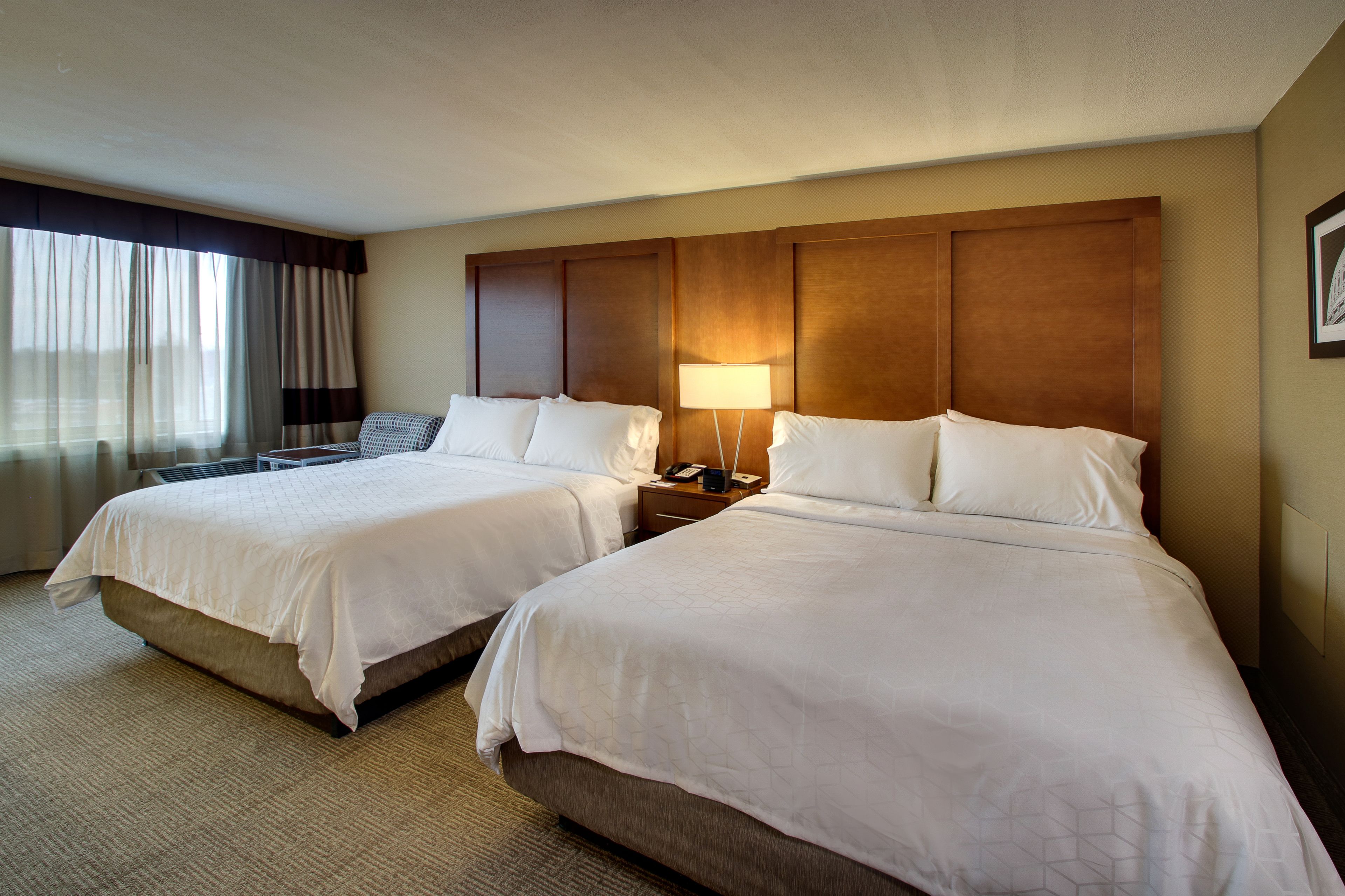 A hotel room with two beds, a nightstand with a lamp, and a large window with curtains.