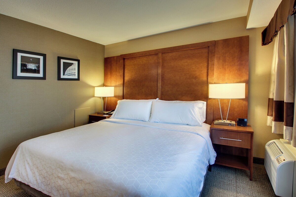 A hotel room with a large bed, two bedside lamps, a nightstand, a heater, and two framed pictures on the wall.