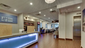 Un espace d’accueil d’hôtel comprenant un comptoir de réception en bois, un réfrigérateur de la marque Coca-Cola et un coin salon avec des chaises et des tables.