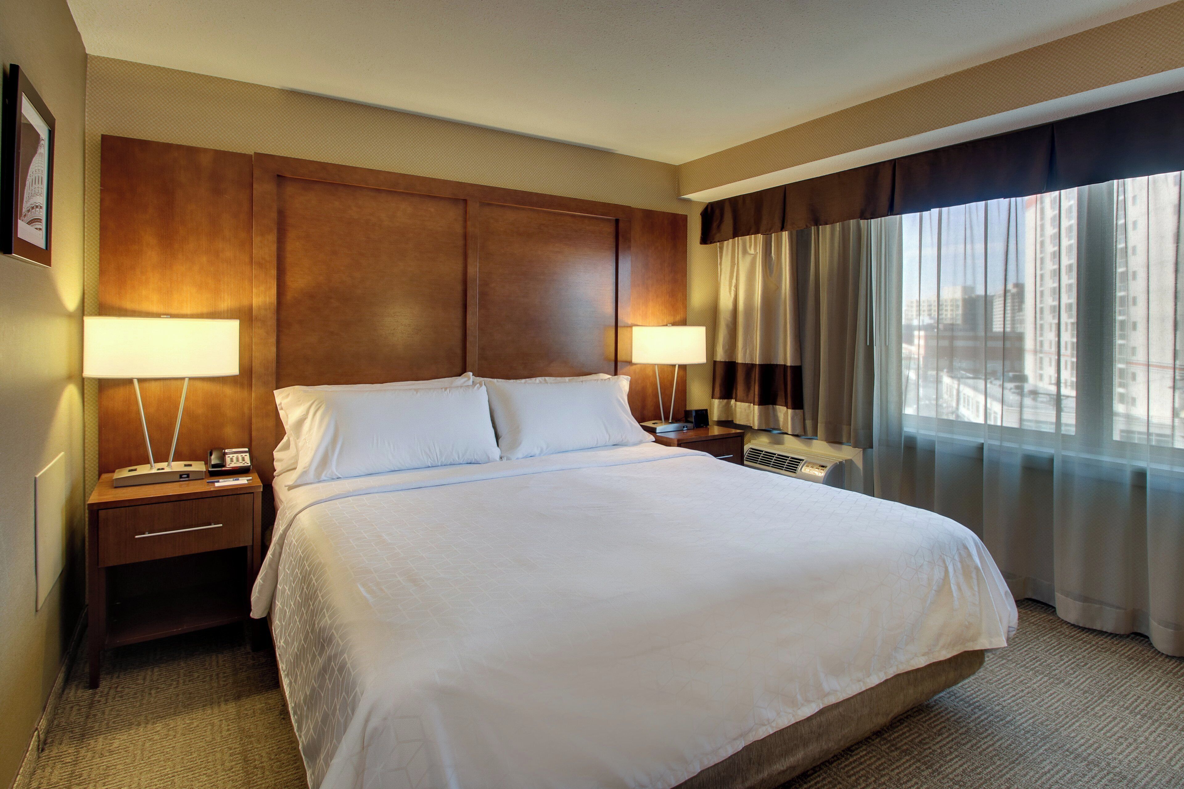 A hotel room with a large bed, two bedside tables with lamps, a window with curtains, and a wall-mounted air conditioning unit.