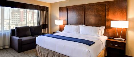A hotel room with a large bed, a sofa, two bedside lamps, and a view of buildings through the window.