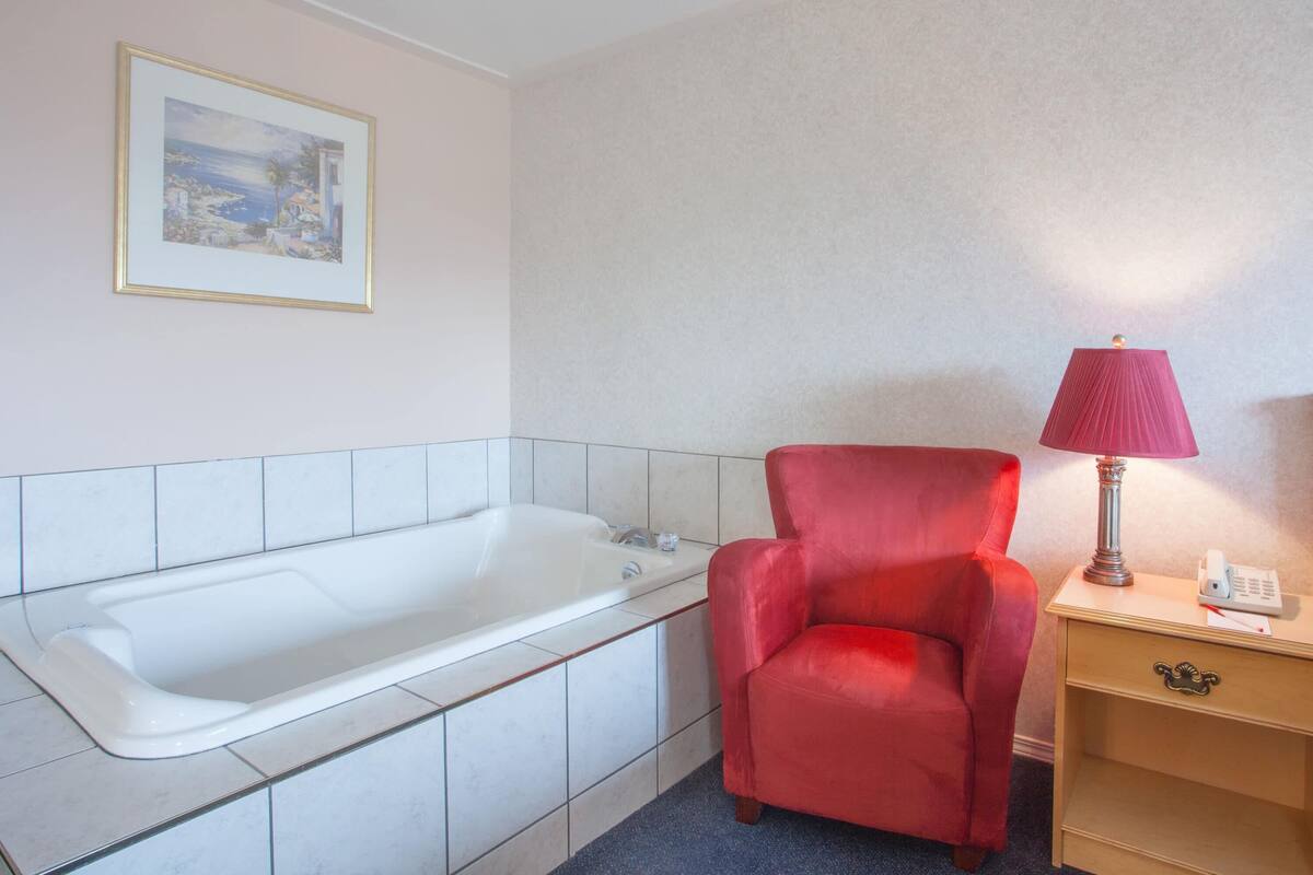 Ein Badezimmer mit einer weißen Badewanne, einem roten Sessel, einem Nachttisch mit Lampe und einem gerahmten Bild an der Wand.