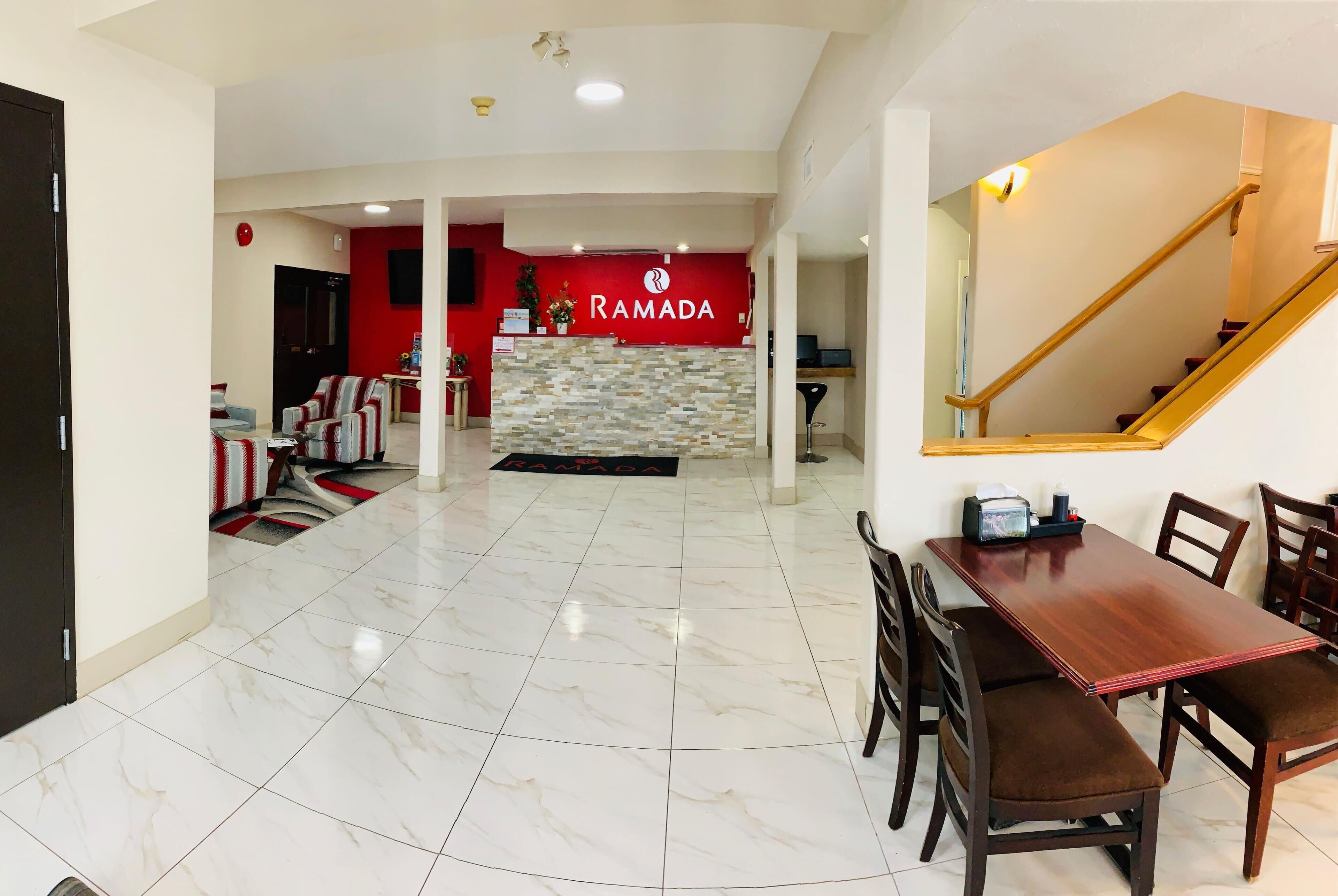 Amplia sala de estar con una mesa y sillas de comedor, una zona de cocina con una pared roja y un cartel que dice "RAMADA", y una escalera con barandillas de madera.