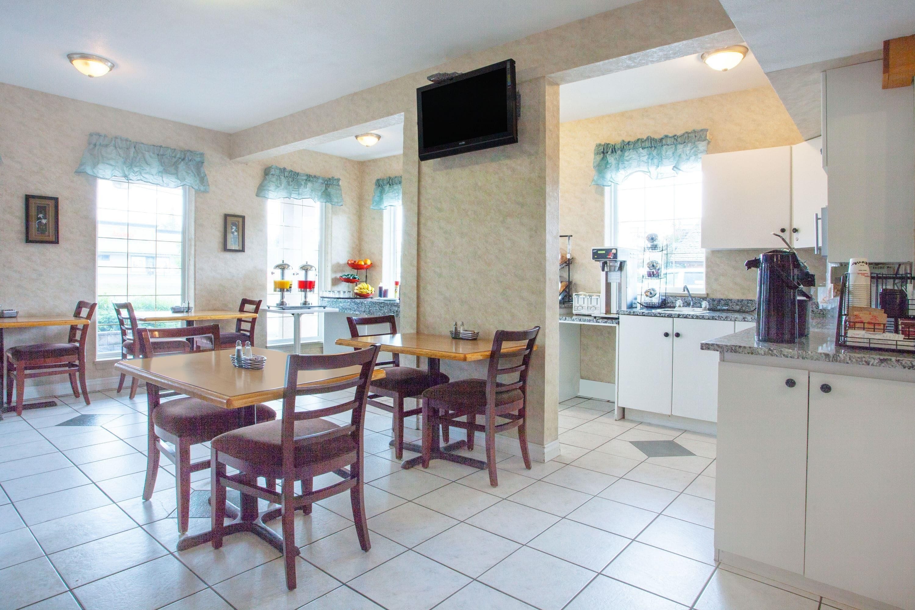 Uma sala de jantar com mesas e cadeiras de madeira, uma cozinha com armários brancos e uma televisão fixada na parede.