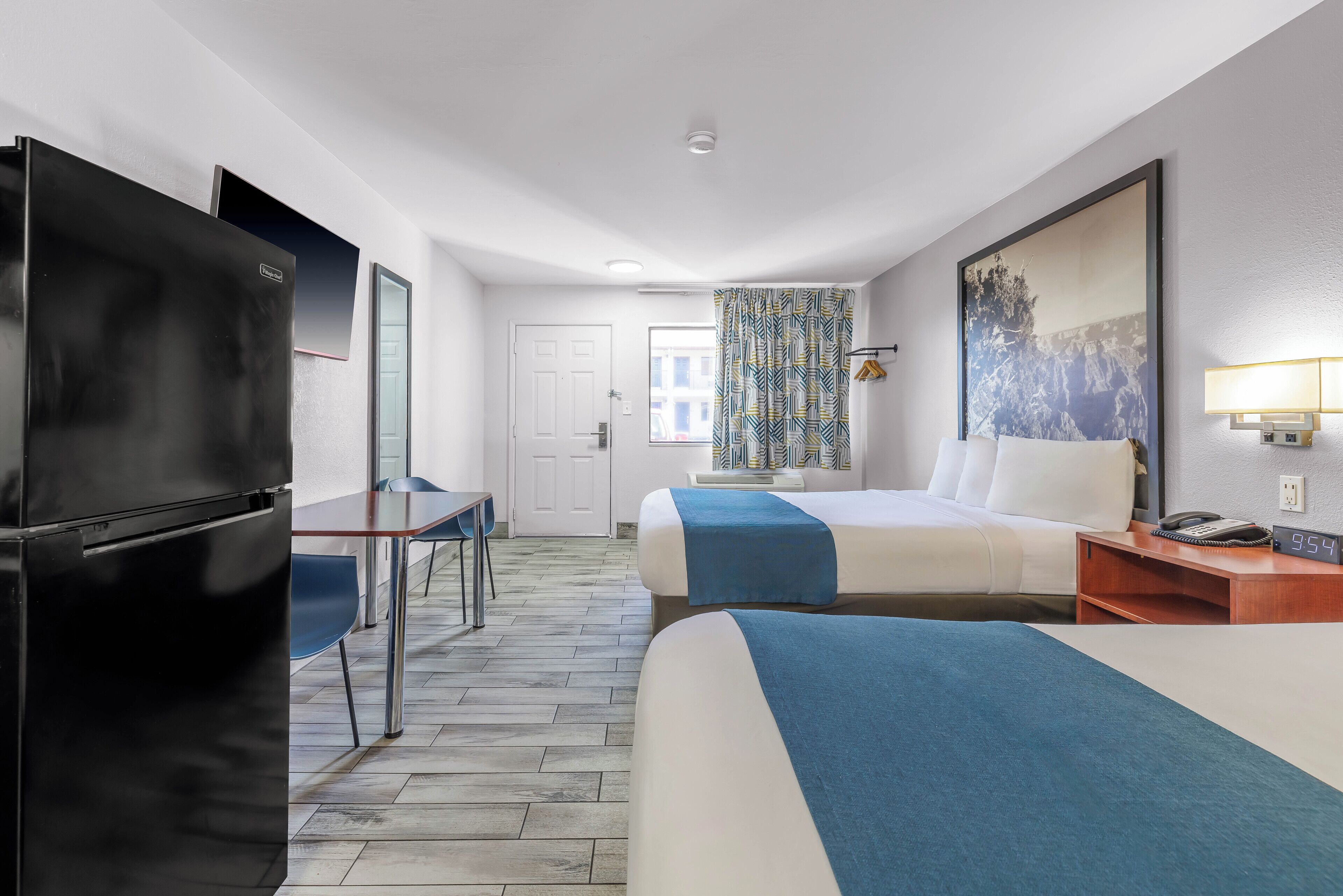 A hotel room with a bed, a desk, a refrigerator, a window with patterned curtains, and a wall-mounted lamp.