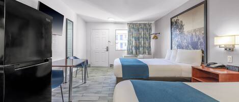 A hotel room with a bed, a desk, a refrigerator, a window with patterned curtains, and a wall-mounted lamp.