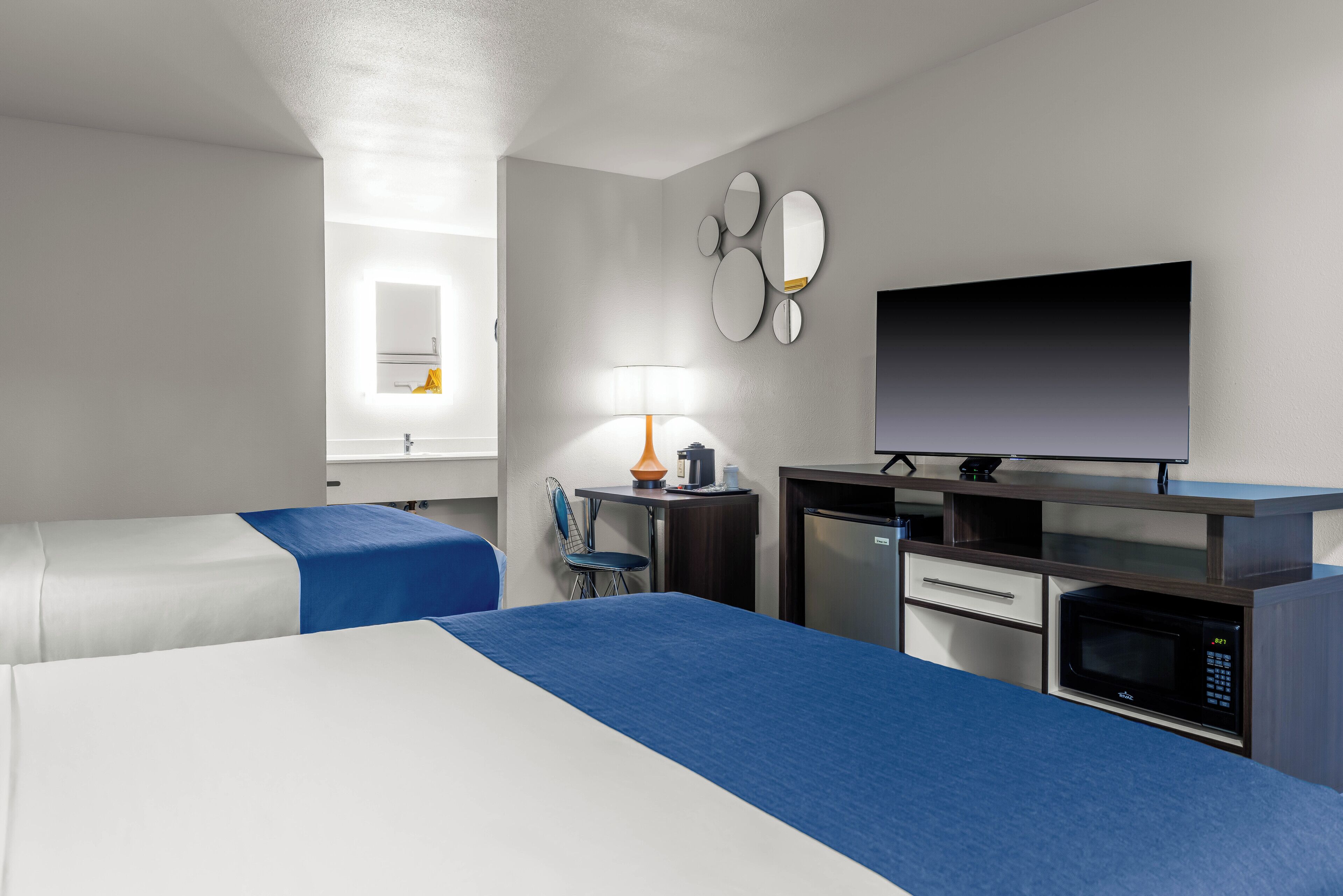 A hotel room with two beds, a desk with a microwave, a TV, a lamp, and a small refrigerator.