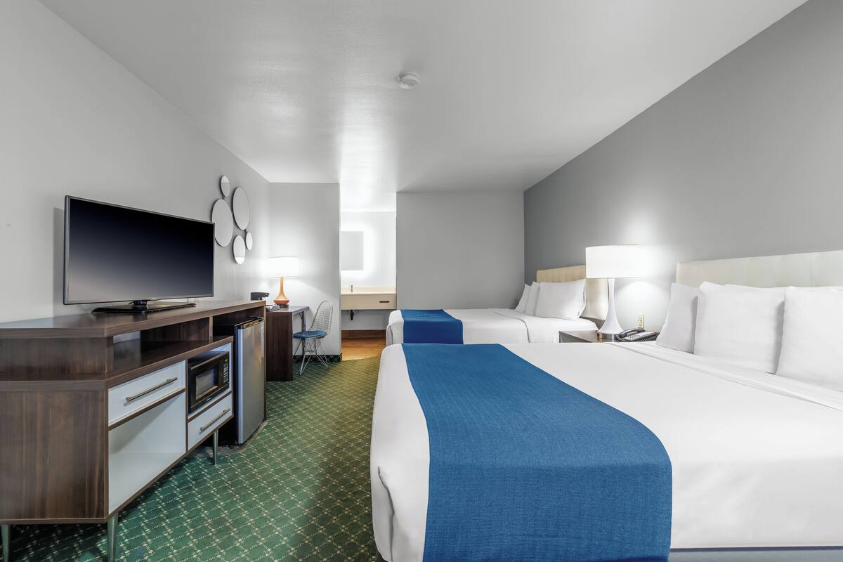 A hotel room with a bed, a desk with a TV, a microwave, a refrigerator, and a chair.