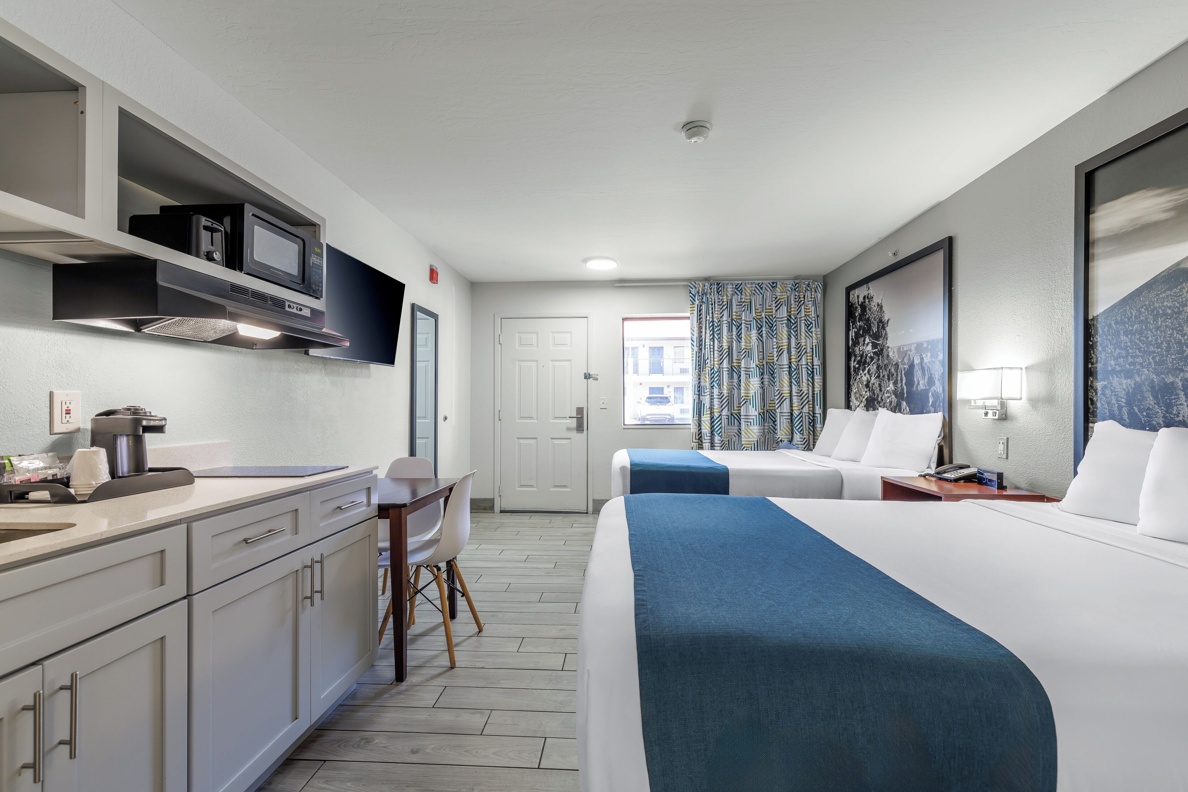 A hotel room with a kitchenette, a bed with a blue blanket, a desk with a chair, and a window with patterned curtains.