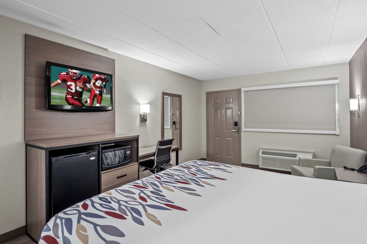 A hotel room with a large bed, a TV mounted on the wall, a desk with a chair, a small fridge, and a window with blinds.
