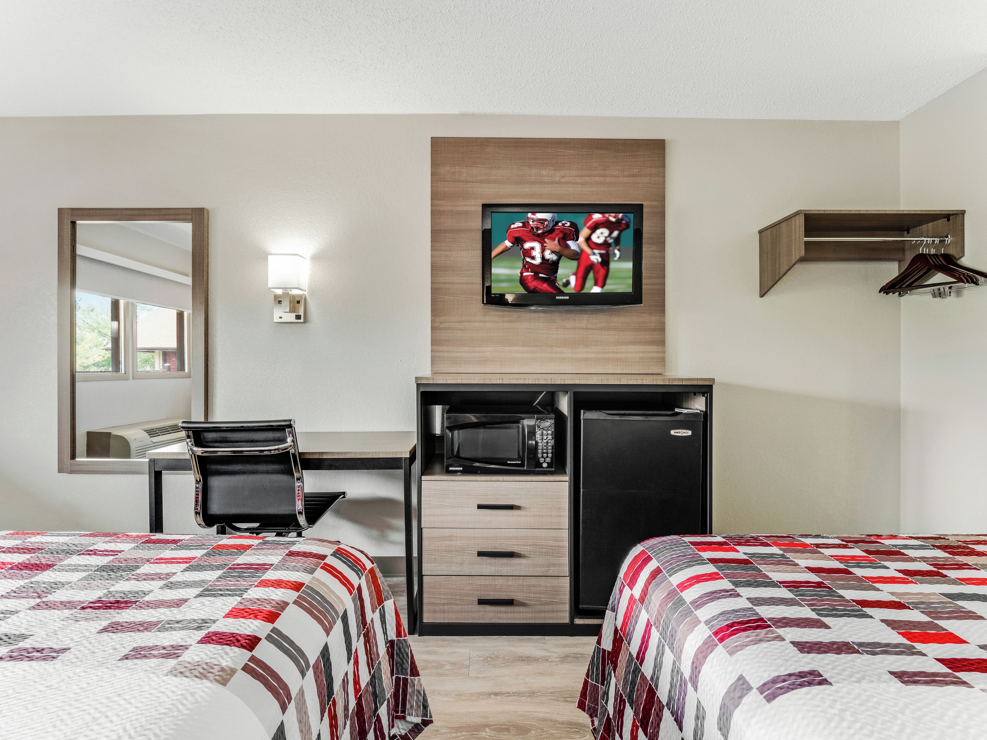 A hotel room with two beds, a desk with a computer, a microwave, and a refrigerator. There is a TV mounted on the wall displaying a sports game.