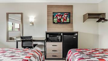 A hotel room with two beds, a desk with a computer, a microwave, and a refrigerator. There is a TV mounted on the wall displaying a sports game.