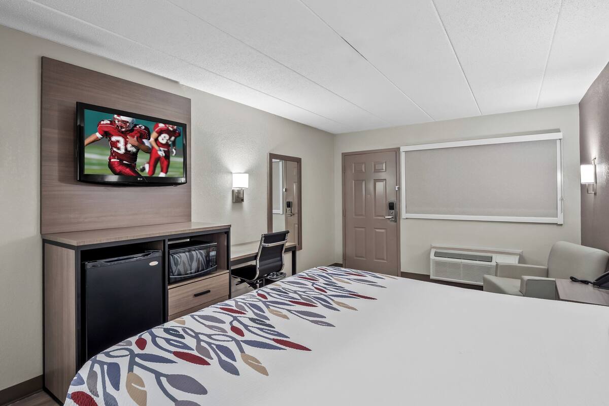 A hotel room with a large bed, a TV mounted on the wall, a desk with a chair, a small fridge, and a window with blinds.