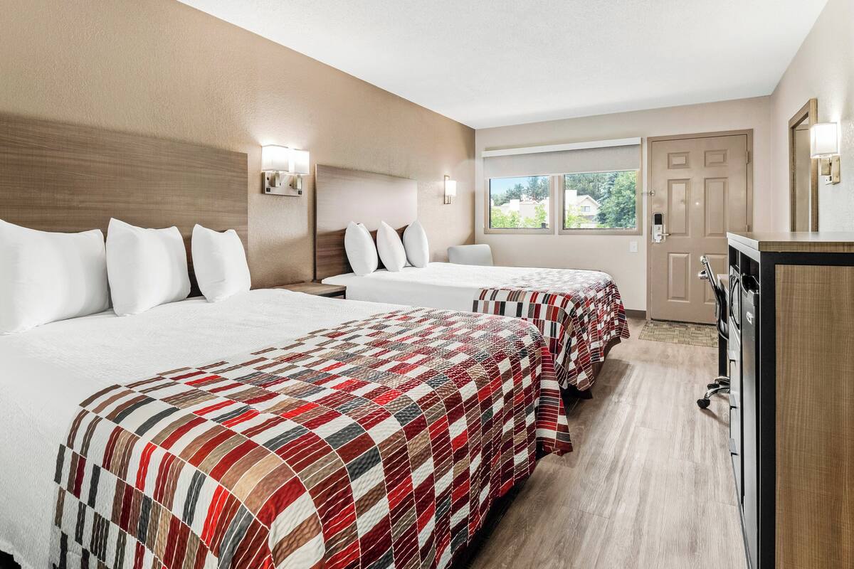 A hotel room with two beds, a desk, a dresser, a window with a view, and a door.