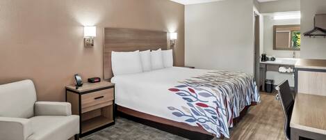 A hotel room with a bed, bedside table, chair, desk, and a small kitchenette area.