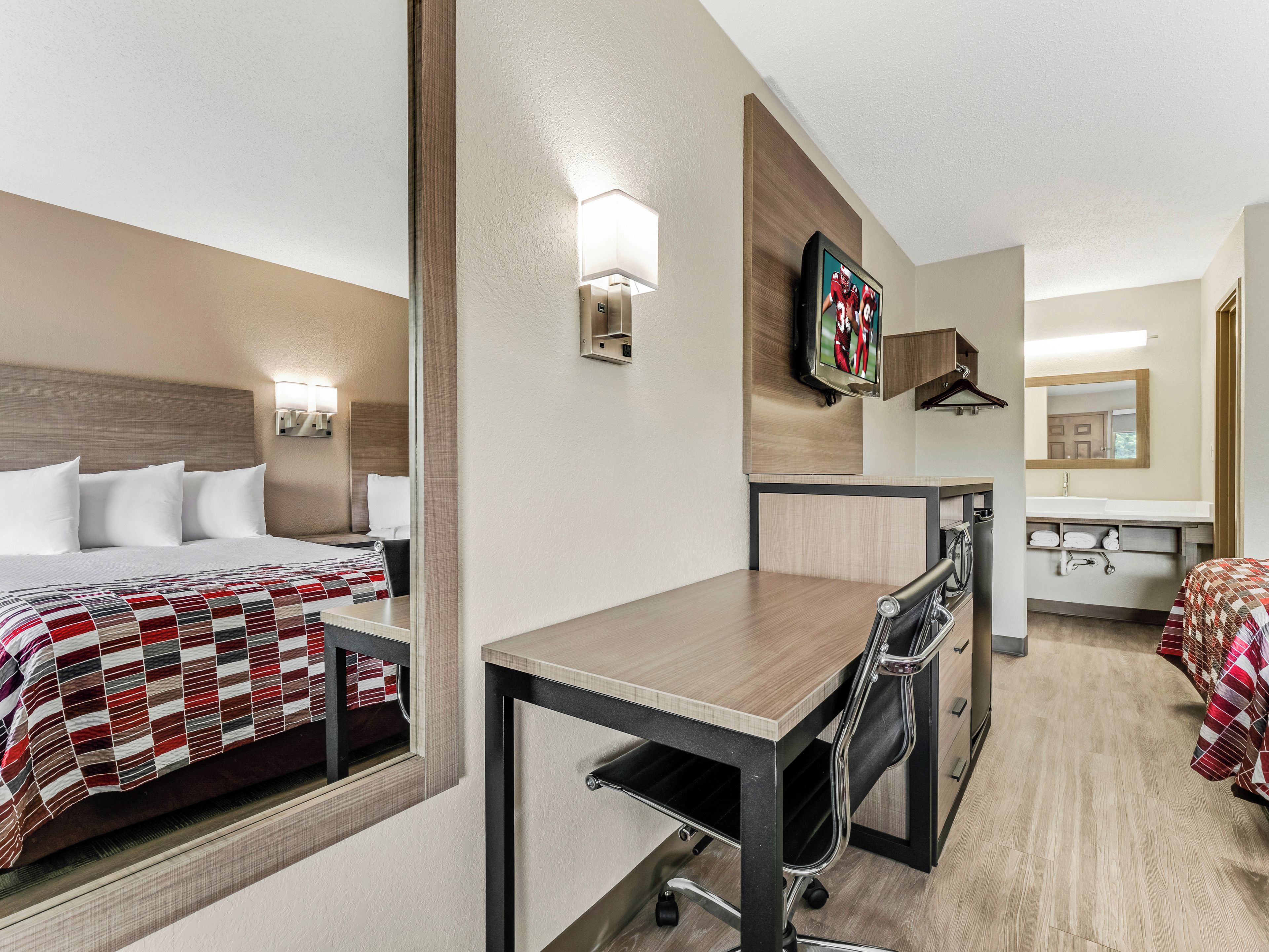 Una habitación de hotel con cama, escritorio, silla, espejo, televisor y baño.