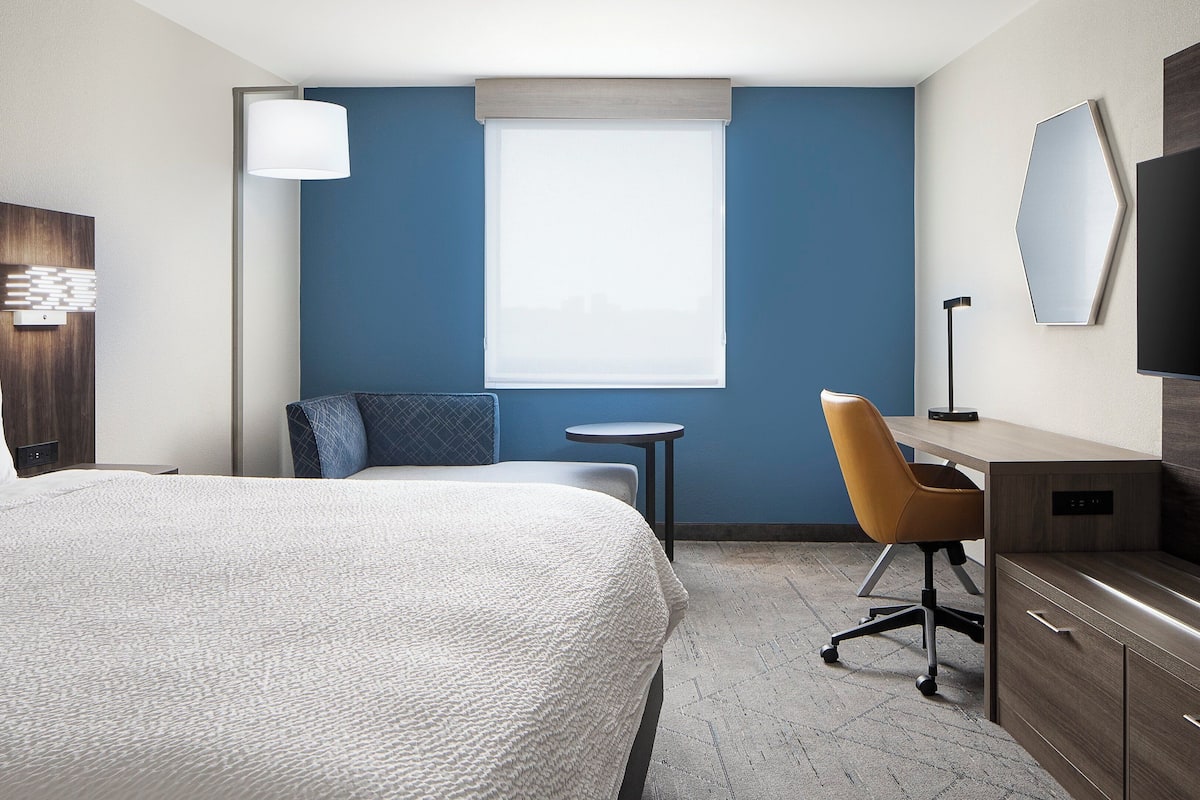 A hotel room with a large bed, a desk with a chair, a small table, a TV, and a window with blinds.