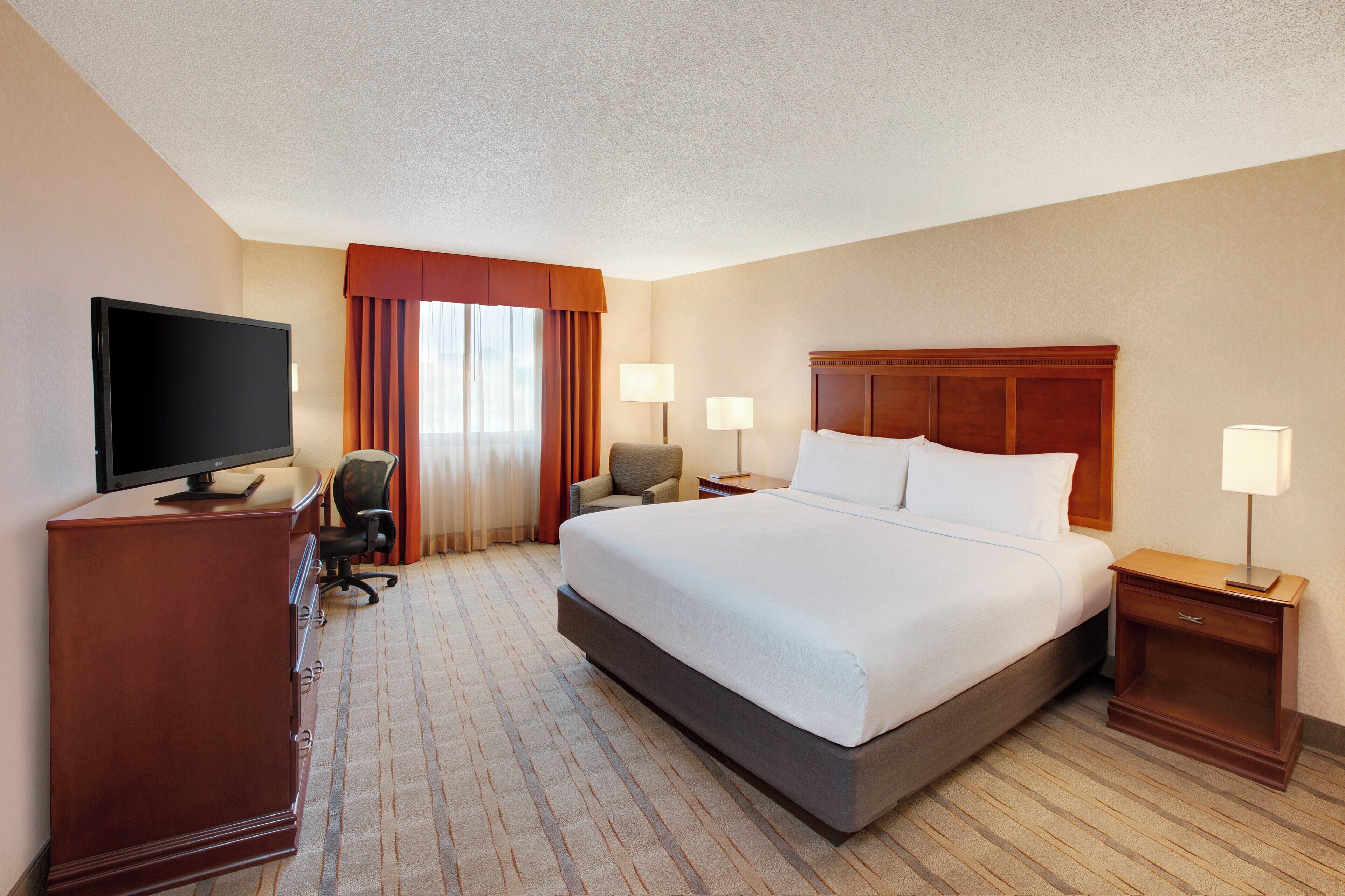 A hotel room with a large bed, a desk with a flat-screen TV, two bedside lamps, and a chair.