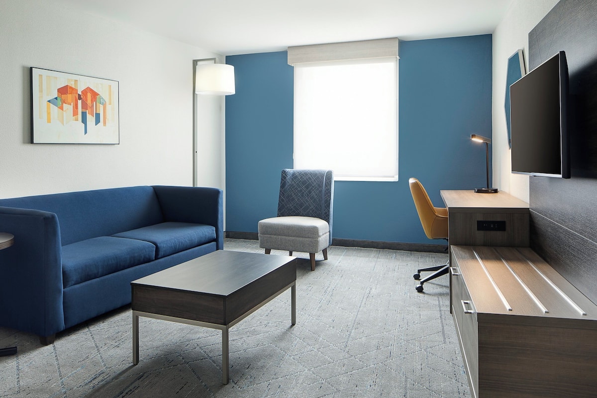 A modern hotel room with a blue sofa, a small table, a chair, a desk with a flat-screen TV, and a painting on the wall.