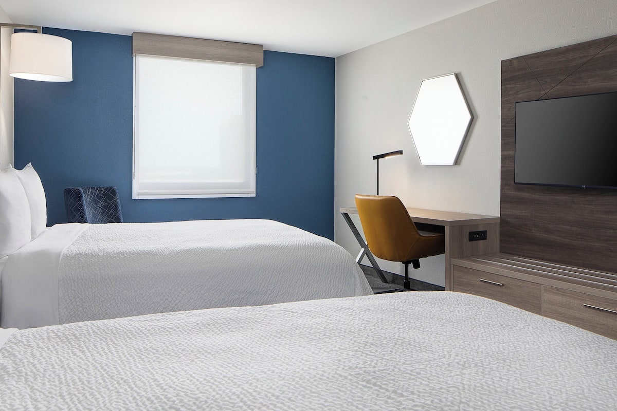 A hotel room with two beds, a desk, a chair, a TV, and a window with blinds.