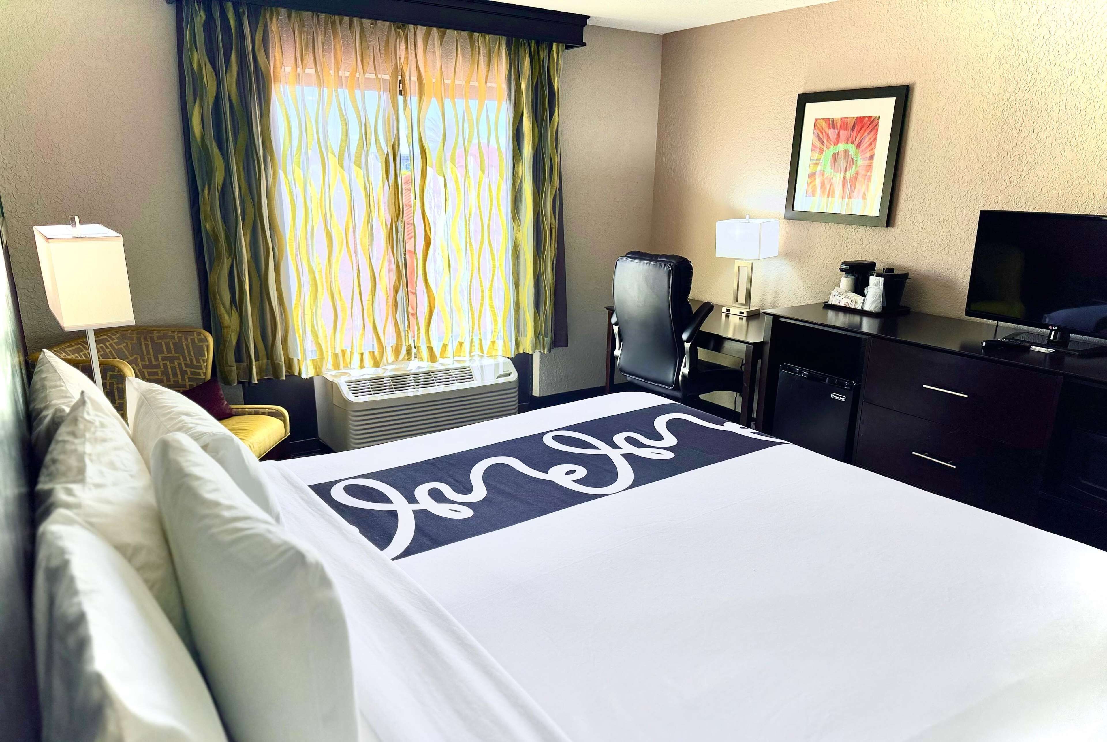 A hotel room with a bed, a desk with a computer, a chair, a TV, and a window with curtains.