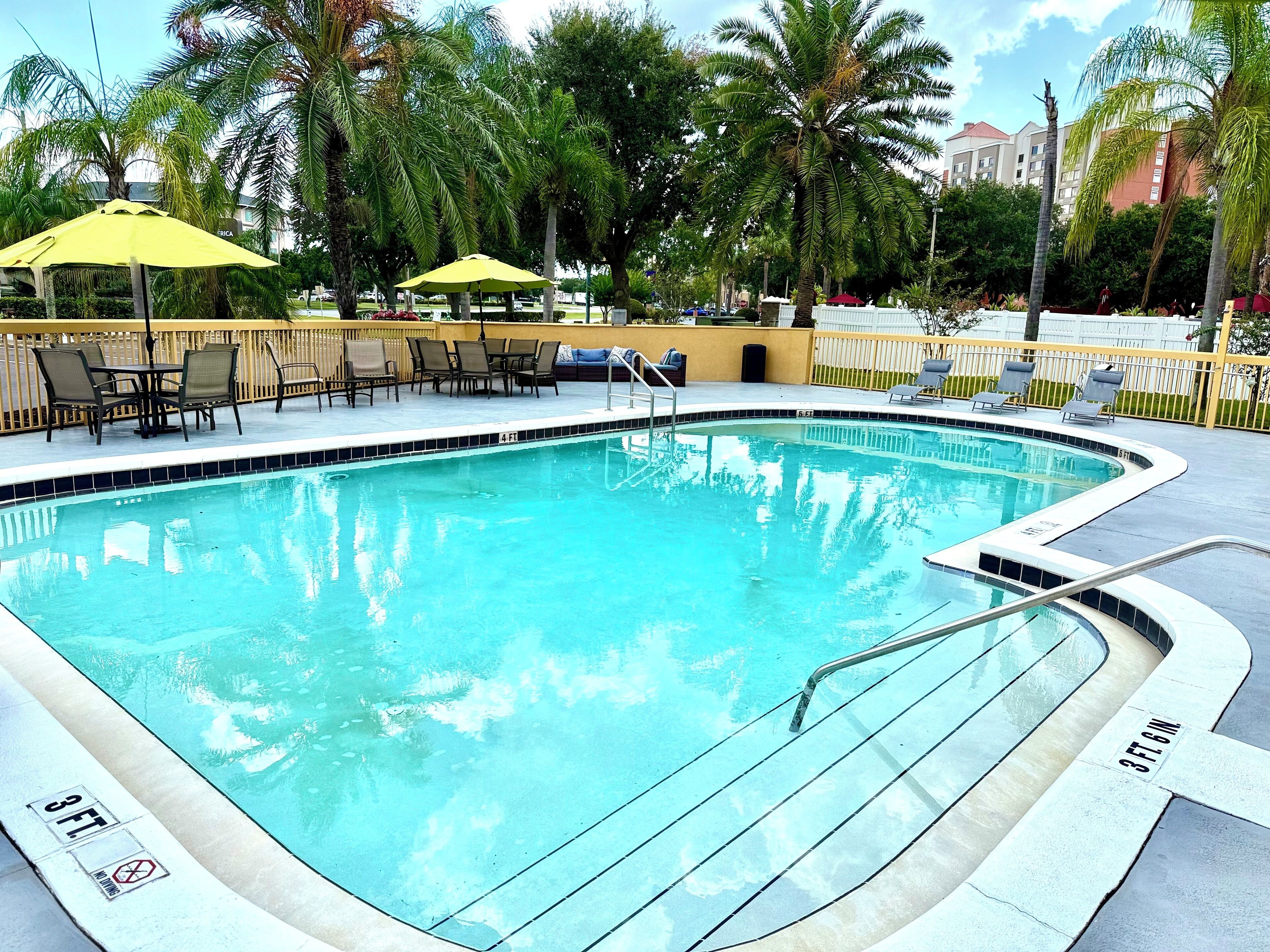 A swimming pool with a 3 ft. depth marker, surrounded by lounge chairs and umbrellas, with palm trees and buildings in the background.