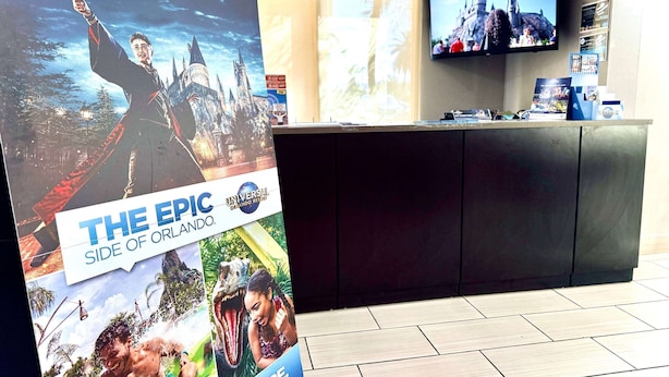 A hotel reception area with a large poster advertising 'The Epic Side of Orlando', a flat-screen TV on the wall, and a reception desk with various items on it. - La Quinta Inn & Suites by Wyndham Orlando Universal area (Orlando)