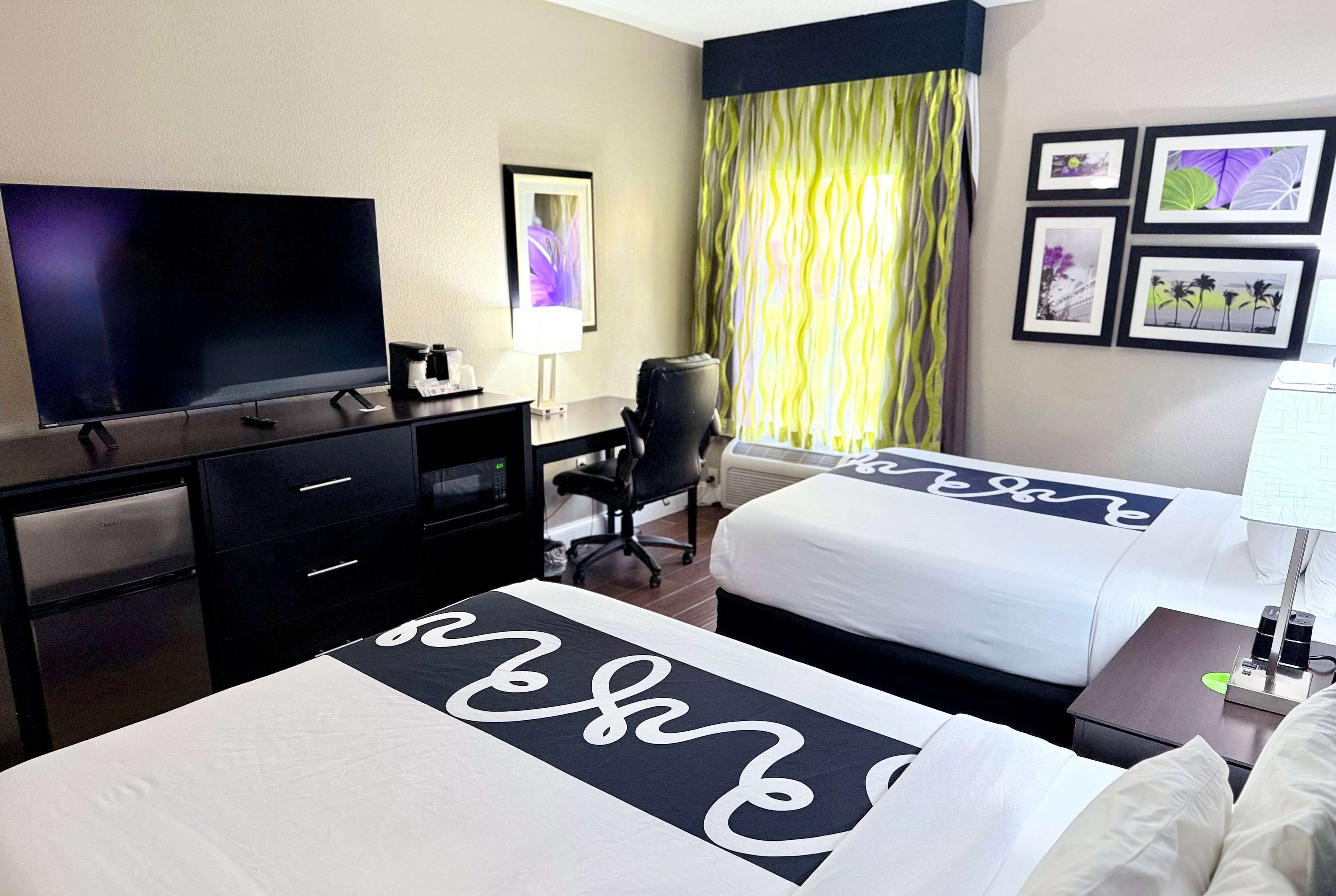 Ein Hotelzimmer mit Fernseher, Schreibtisch, Stuhl, zwei Betten und einem Kunstwerk an der Wand.