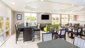A modern dining area with multiple tables and chairs, a flat-screen TV mounted on the wall, and large windows providing natural light. - La Quinta Inn & Suites by Wyndham Orlando Universal area (Orlando)