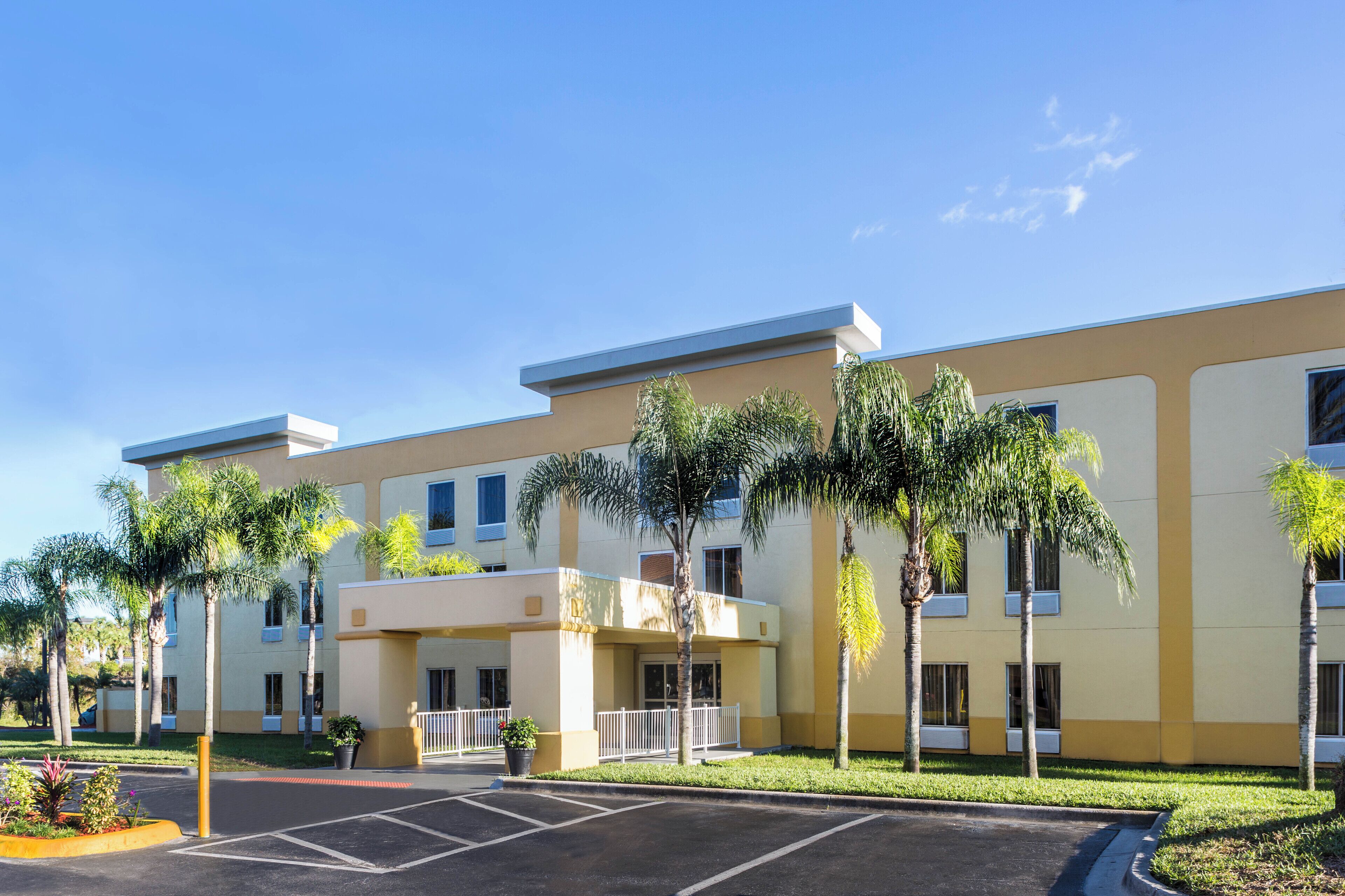 A multi-story hotel with a yellow facade, a parking lot, and palm trees.