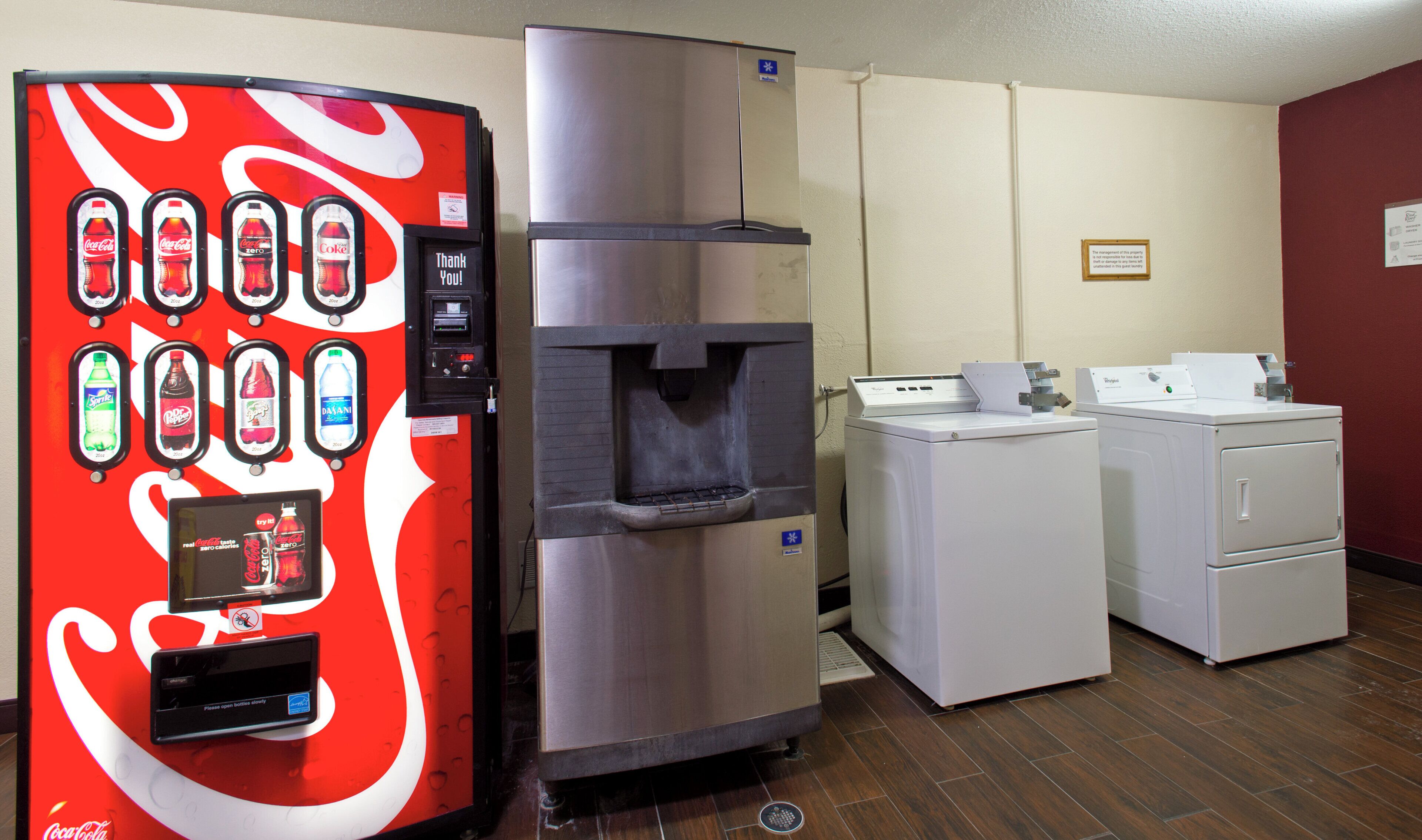 A vending machine with various drink options, a stainless steel refrigerator, and a washing machine in a room with wooden flooring.