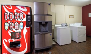 Une machine distributrice proposant diverses boissons, un réfrigérateur en acier inoxydable et un lave-linge, le tout dans une pièce dont le sol est recouvert de parquet.