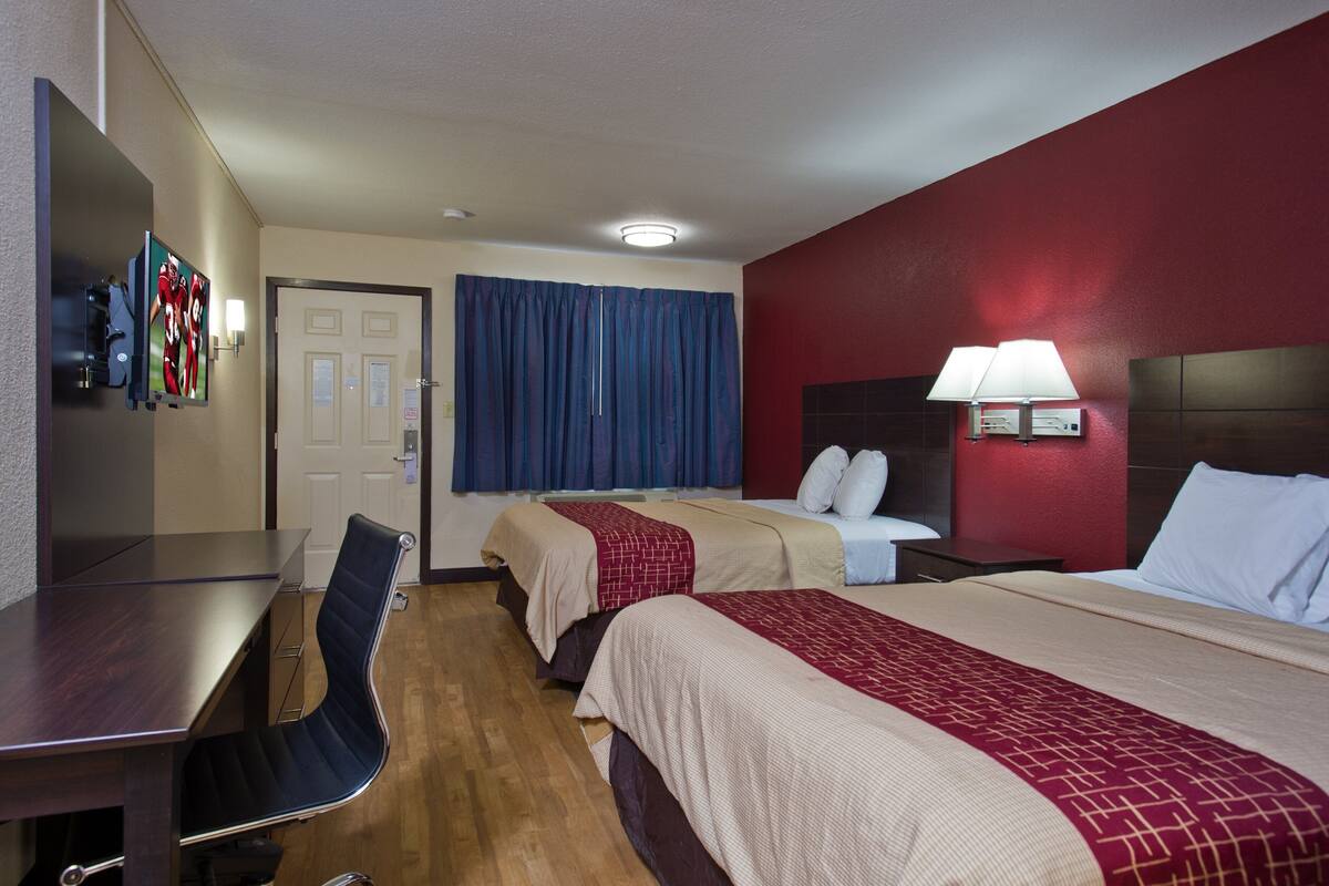 Ein Hotelzimmer mit zwei Betten, einem Schreibtisch, einem Stuhl, einem Fernseher und einer roten Akzentwand.