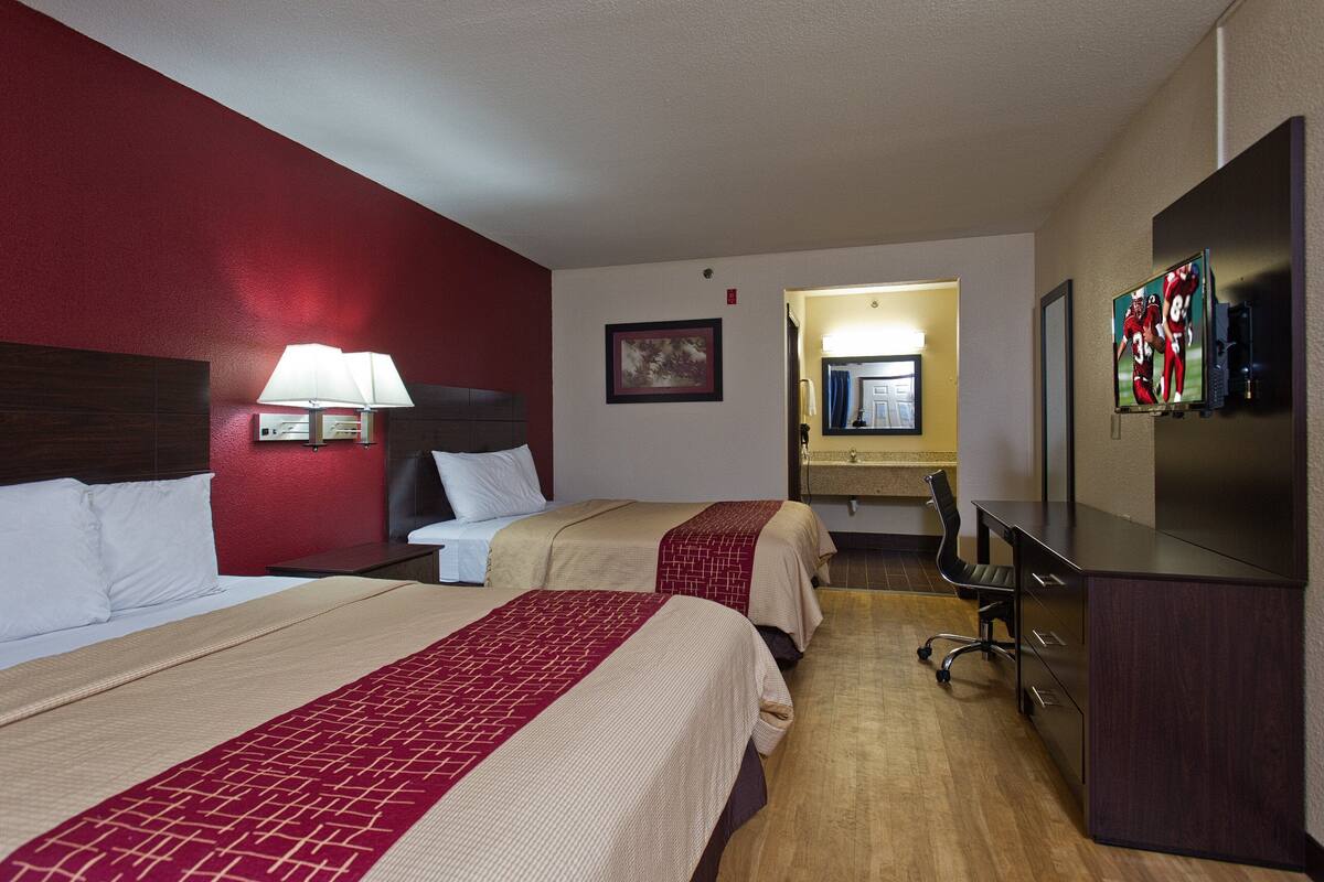 Ein Hotelzimmer mit zwei Betten, einem Schreibtisch, einem Stuhl, einem Fernseher und einem sichtbaren Badezimmer im Hintergrund.