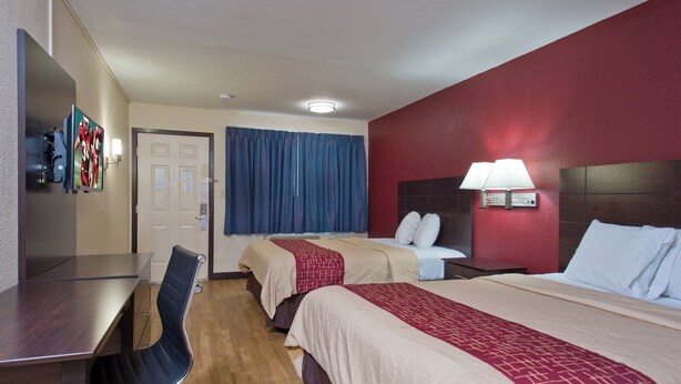 Une chambre d’hôtel avec deux lits, un bureau, une chaise, une télévision et une salle de bain avec un mur rouge et une lampe blanche.