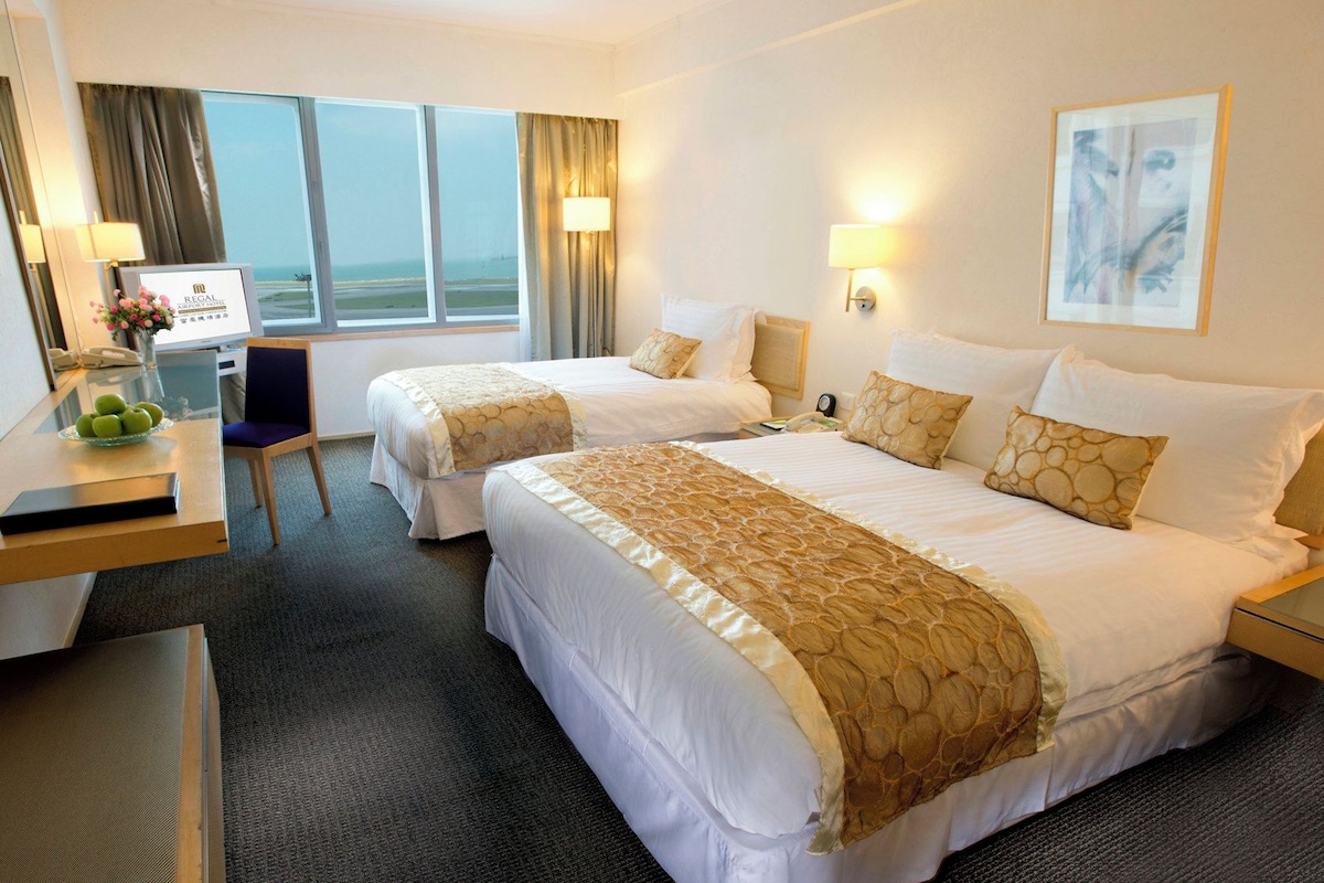 A hotel room with two beds, a desk, a chair, a lamp, and a view of the airport.