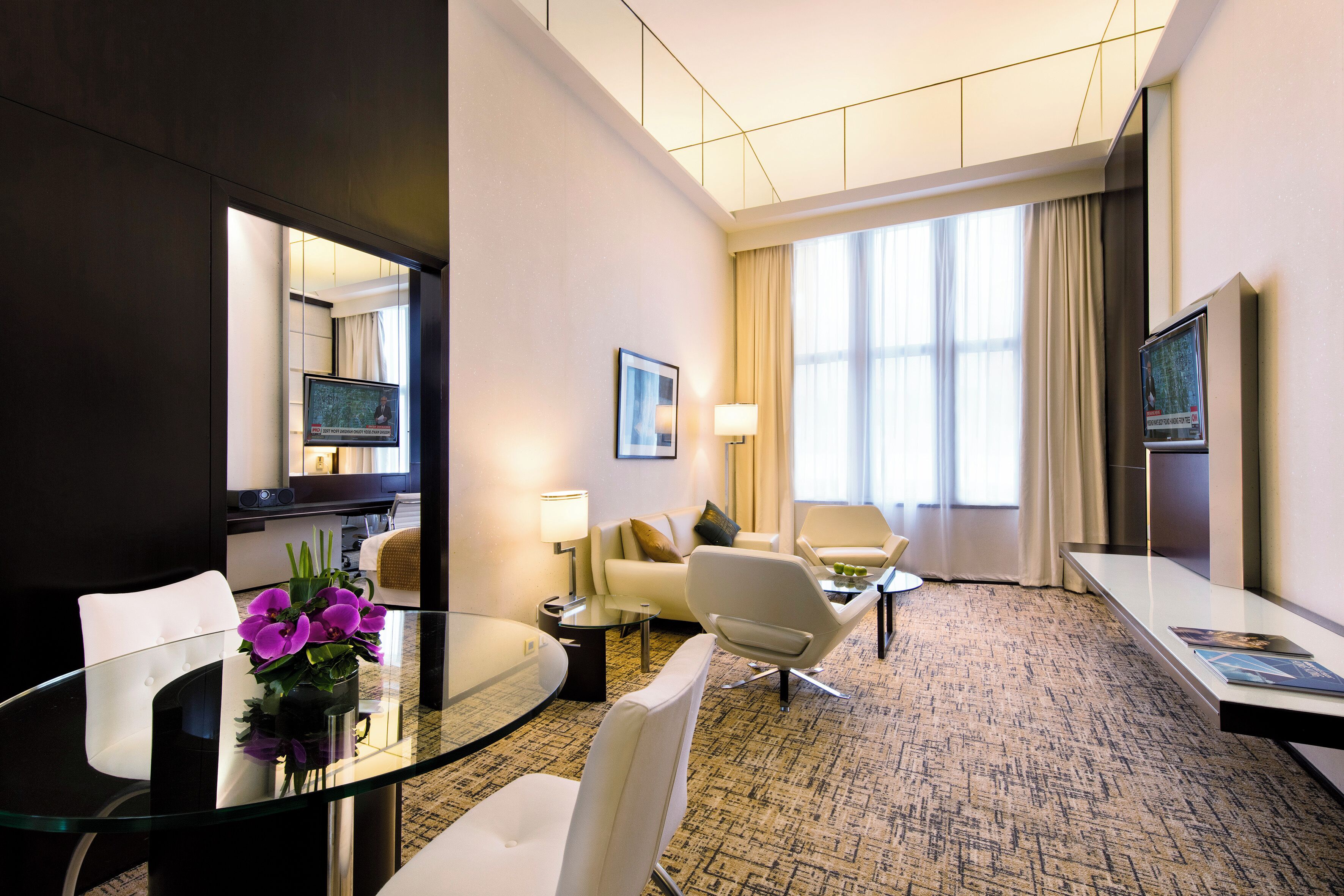 A modern hotel room with a glass table, white chairs, a sofa, a TV, and a large window with curtains.