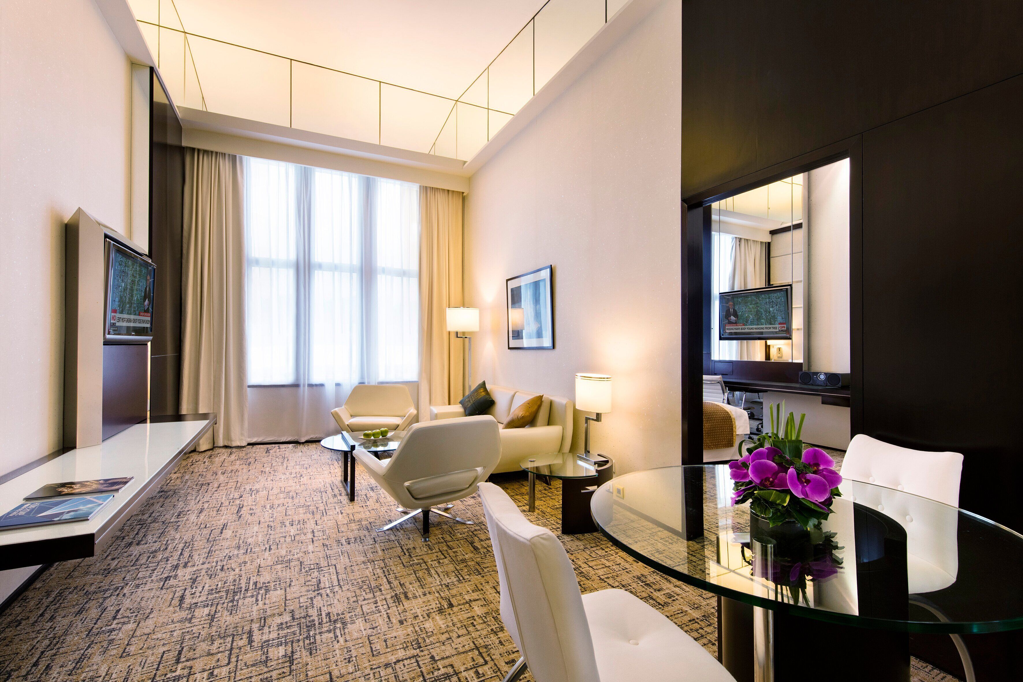 A hotel room with a glass dining table, a sofa, a TV, and a large window with curtains.