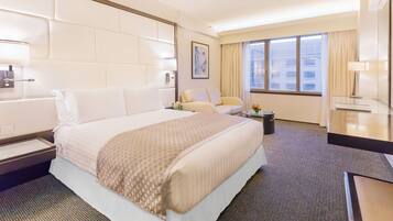 Ein Hotelzimmer mit einem großen Bett, einem Nachttisch, einem Sessel, einem kleinen Tisch mit Blumenvase und einem Fenster mit Vorhängen.