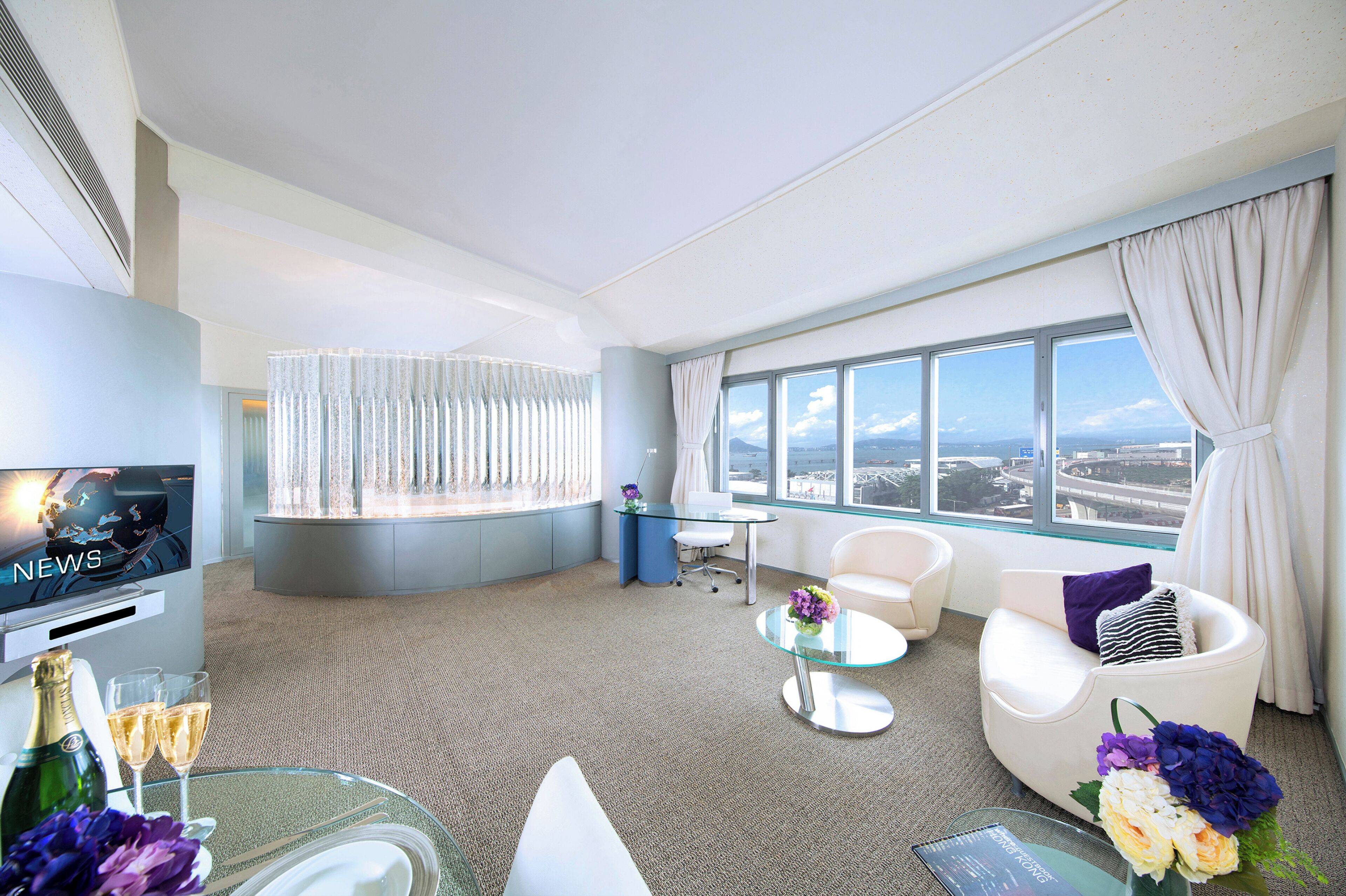 A modern hotel room with a large window, a flat-screen TV, a glass coffee table, and a seating area with a white sofa and armchairs.
