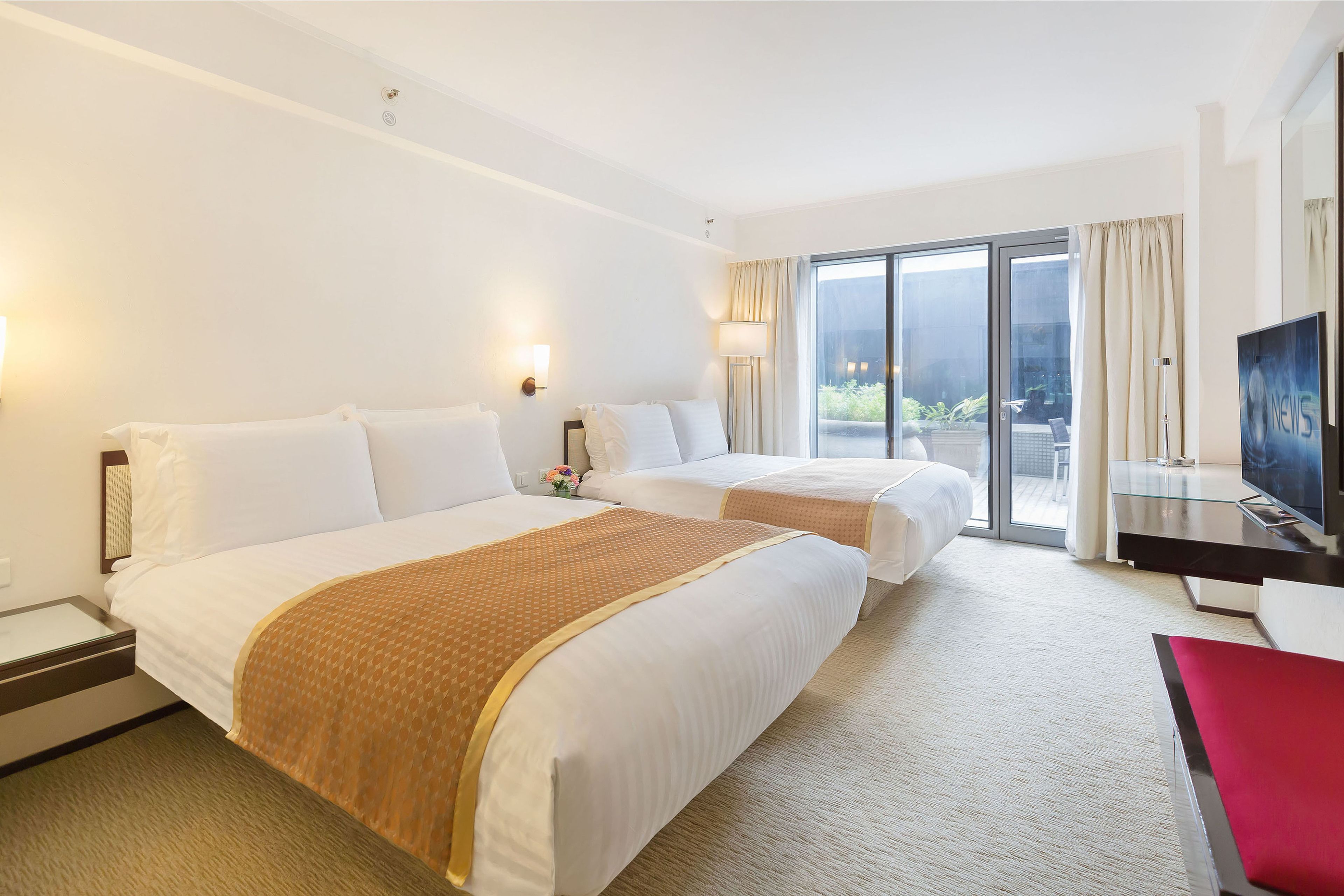 A hotel room with two beds, a TV, a desk, and a balcony with a view.
