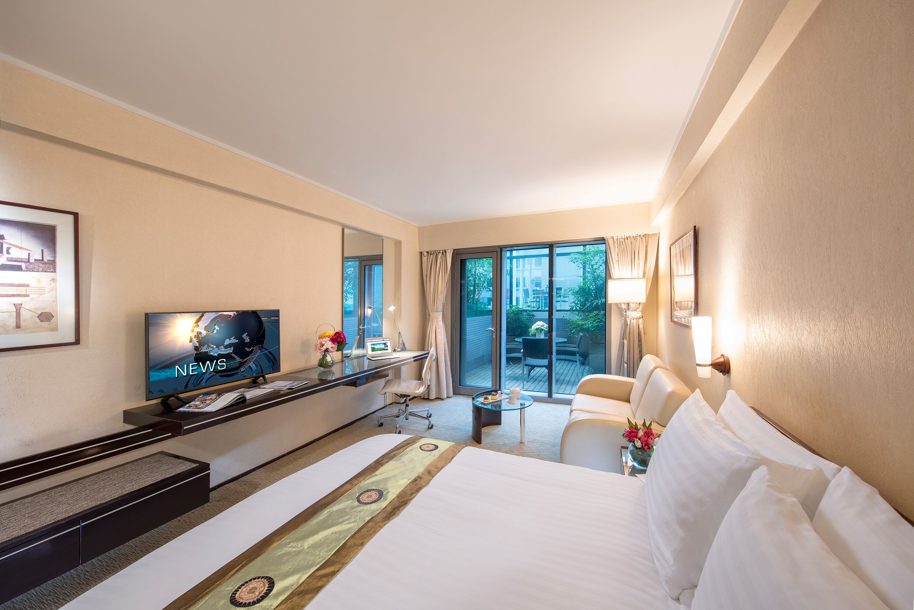 Habitación de hotel con una cama grande, un escritorio con una televisión mostrando 'NEWS', una mesita con un florero y vistas a un balcón con mobiliario exterior.
