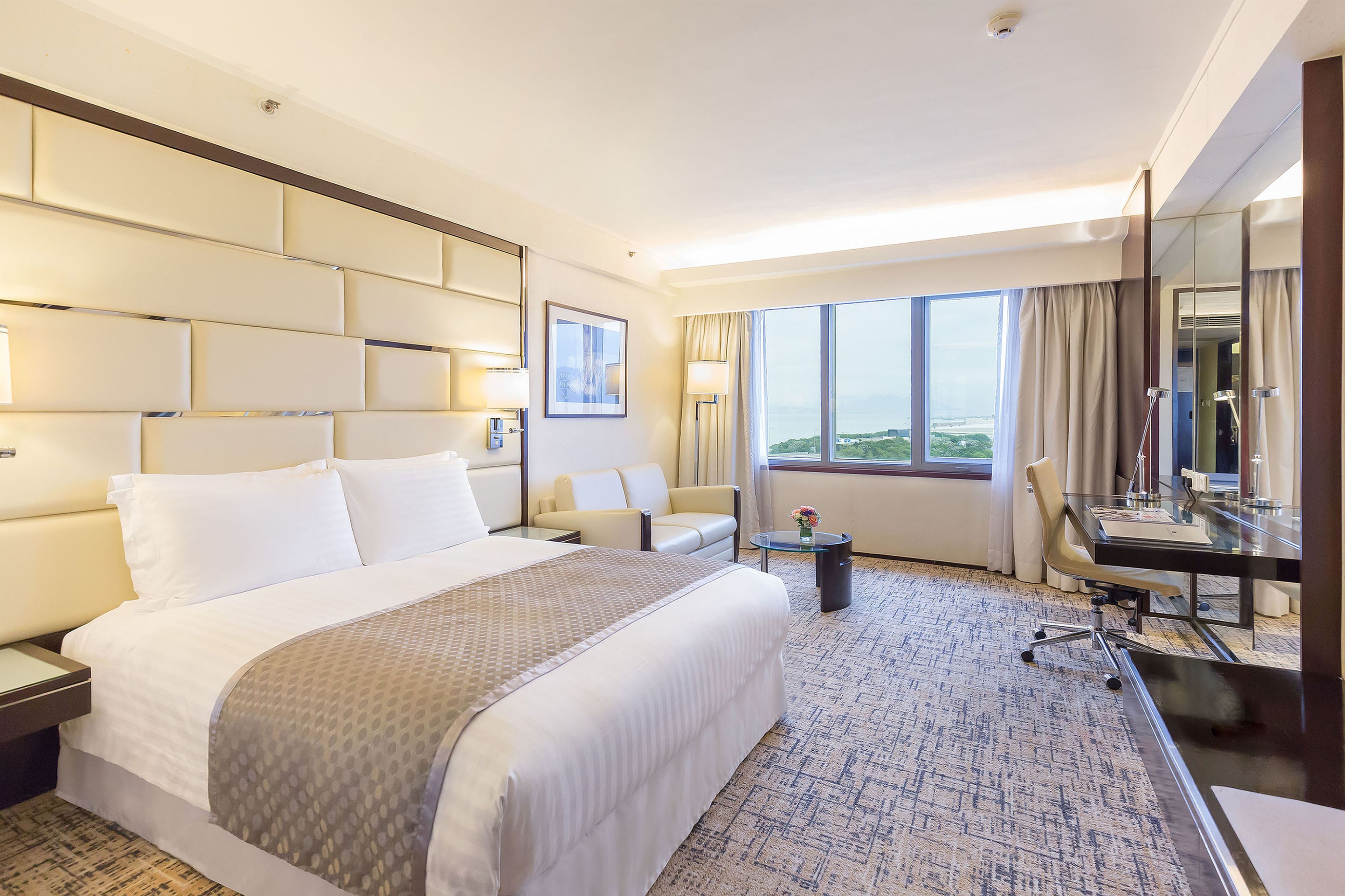 A hotel room with a large bed, a desk, a chair, a small table, and a large window offering a view.