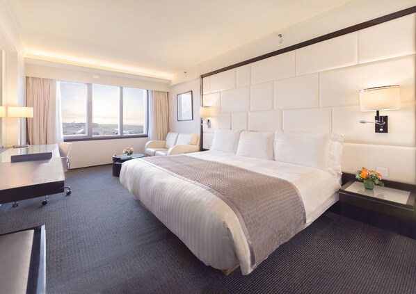 A hotel room with a large bed, a desk, a chair, a small table with flowers, and a window with curtains. - Regal Airport Hotel (Chek Lap Kok)
