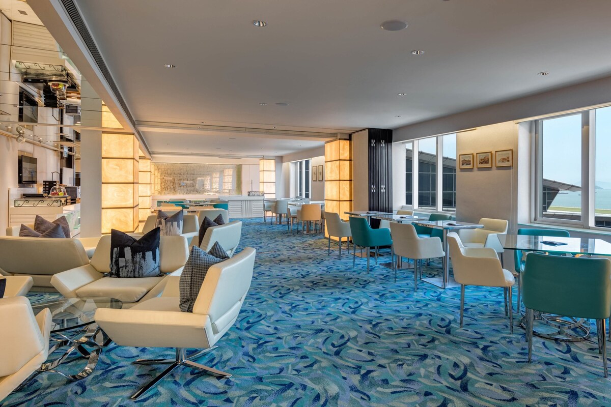 Eine moderne Hotelhalle mit einem blau gemusterten Teppich, Sitzbereichen, einer Bar und großen Fenstern mit Blick auf das Meer.