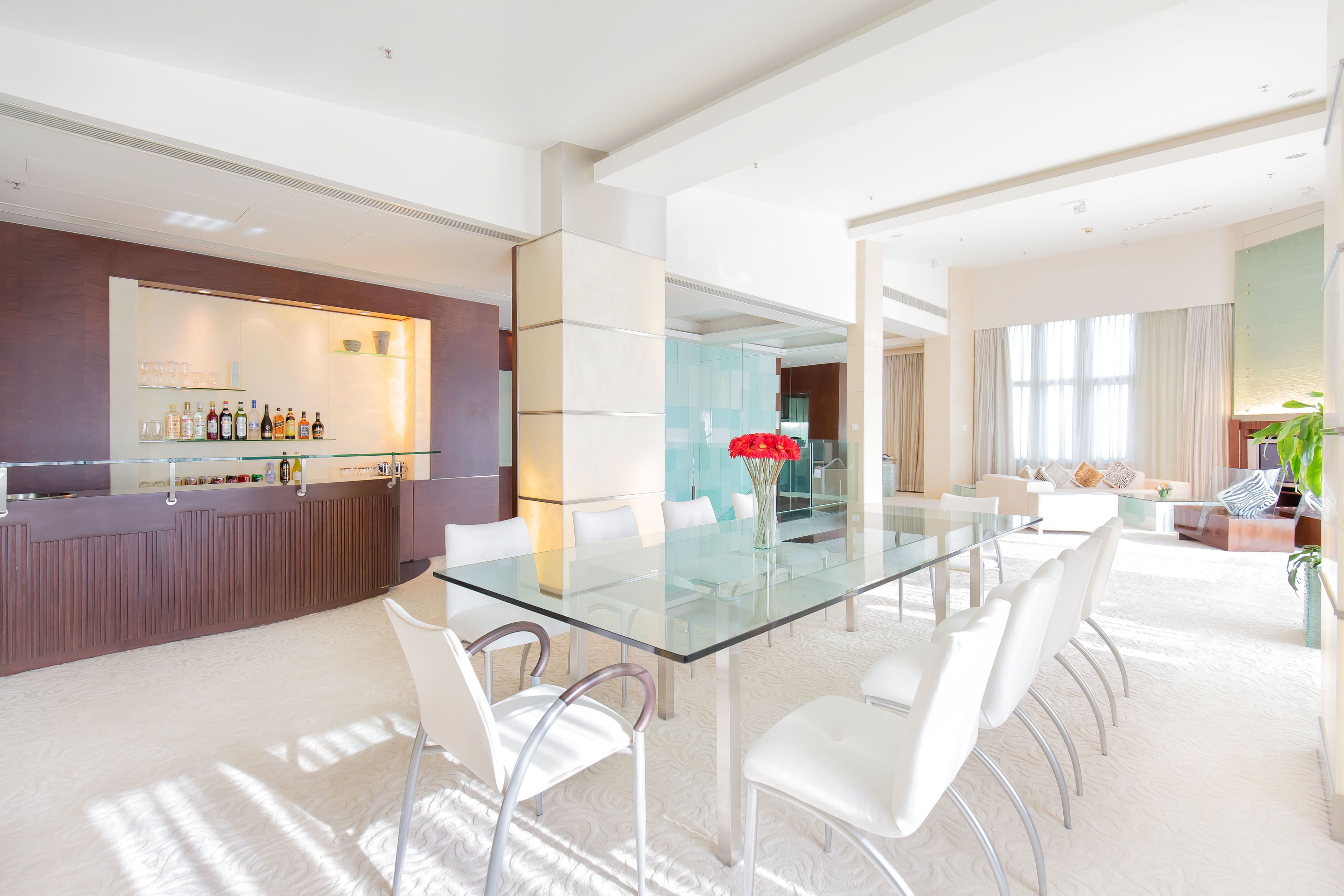 A modern dining area with a glass table and white chairs, a bar with bottles, and a living area with a sofa and curtains.