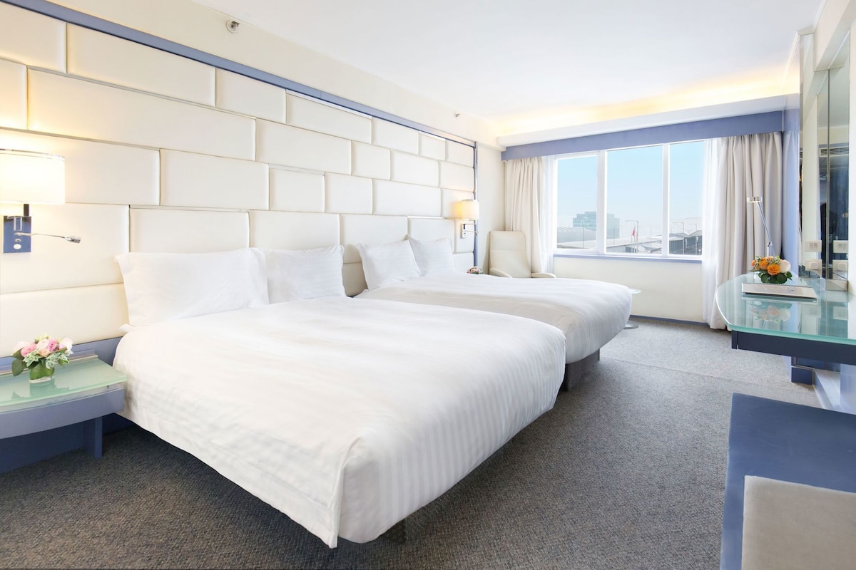 A hotel room with two beds, a desk, a chair, a window with curtains, and a city view.