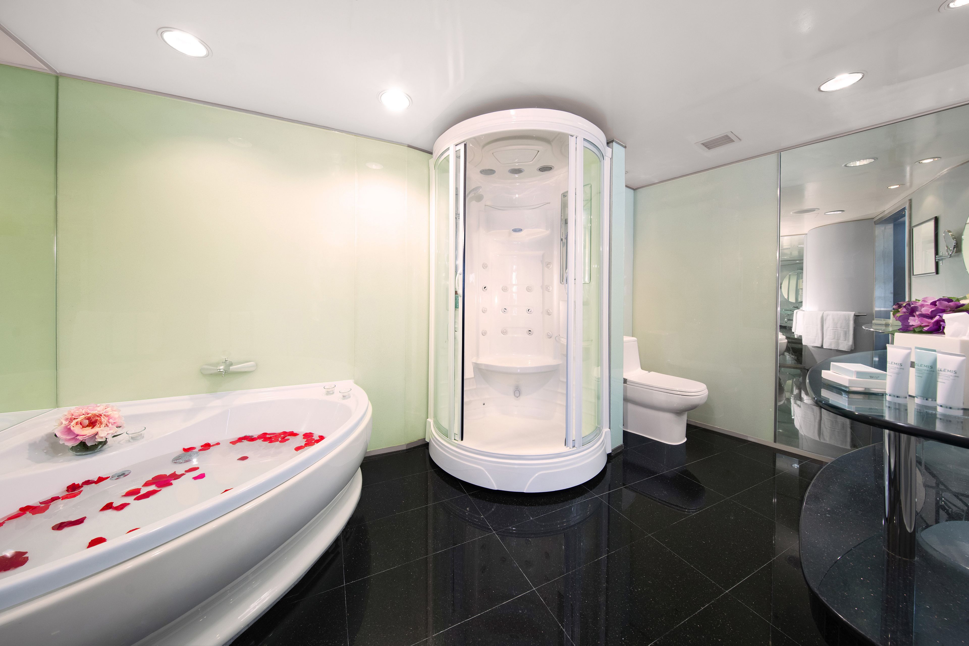A modern bathroom with a glass-enclosed shower, a large bathtub with rose petals, and a glass vanity.