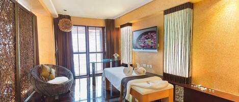 Habitación de hotel con cama, zona de estar con una silla, mesa de masajes, ventana grande con cortinas y televisor instalado en la pared.
